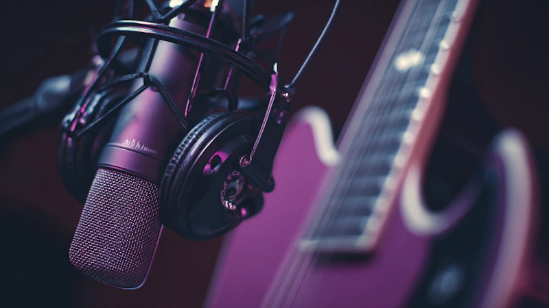 A close-up of a studio condenser microphone with headphones resting on it, ready for a recording session. An electric guitar is softly out of focus in the background, and the entire scene is bathed in atmospheric purple and pink light.
