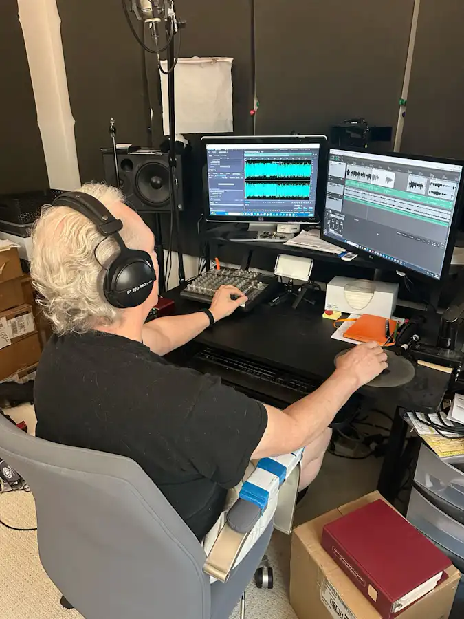Ken Theriot in a black shirt sits in a studio chair with headphones, using dual monitors to edit audio waveforms at his recording studio.