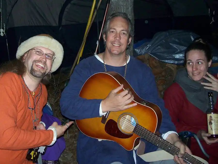 Ken Theriot sits between friends at a nighttime campsite, smiling and playing an acoustic guitar, with one man in a fur hat and a woman holding a bottle.