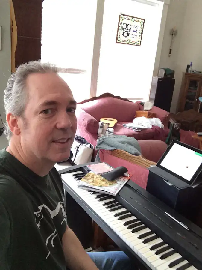 Ken Theriot sits at a digital piano in his living room, composing music, smiling at the camera with a laptop and sheet music on the keyboard.