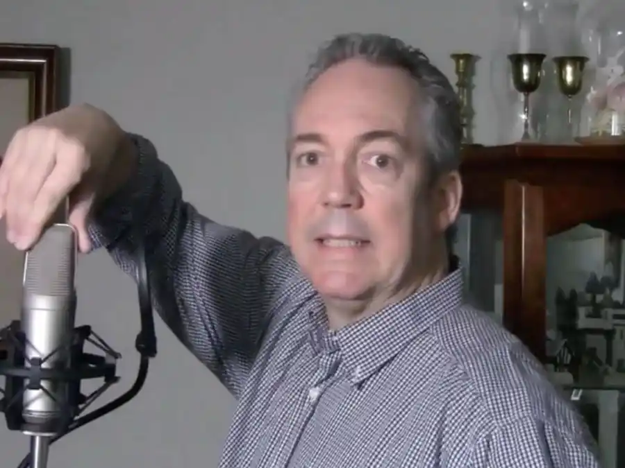 Ken Theriot in a checkered shirt adjusts a studio microphone in a home recording space with glassware displayed behind him.