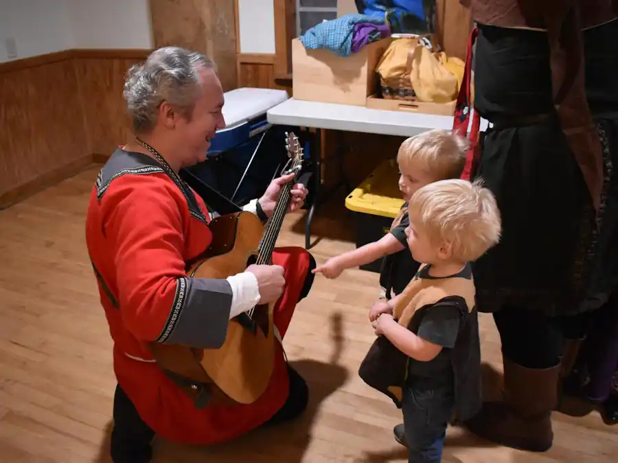 Ken Theriot kneeling in a medieval red tunic demonstrates his guitar to two young children in a wooden hall, showcasing musical curiosity.