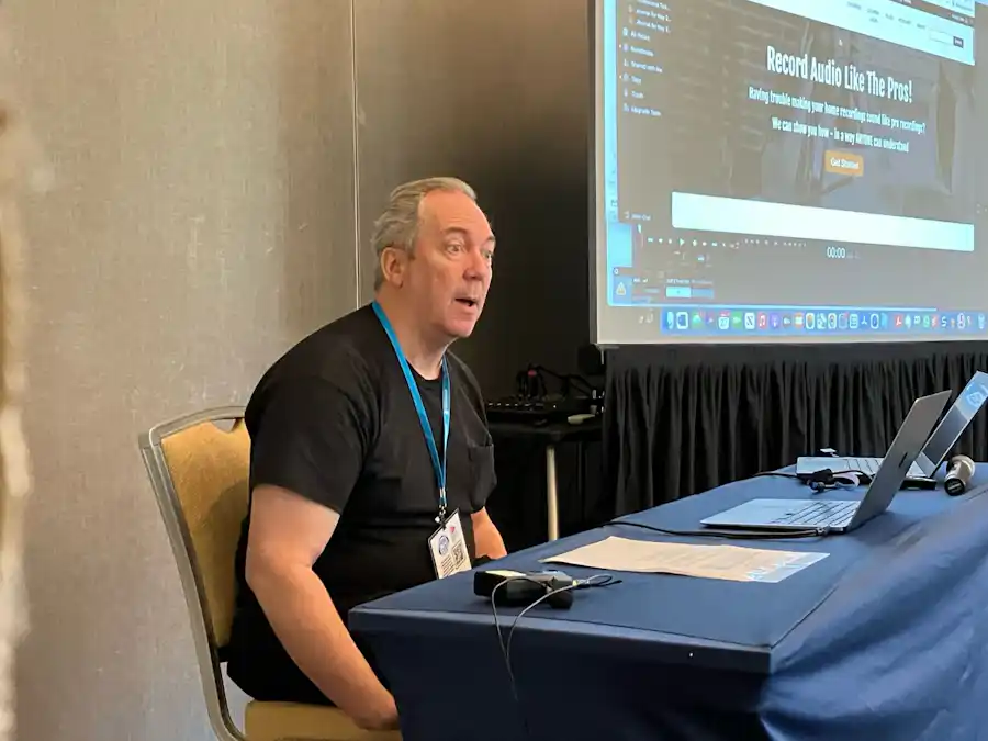 Ken Theriot gives a lecture at a conference table with a laptop, speaking in front of a projected presentation about recording audio.