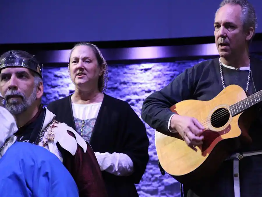 Ken Theriot plays acoustic guitar and sings on stage alongside Lisa Theriot and other performers in medieval costumes under blue lighting.