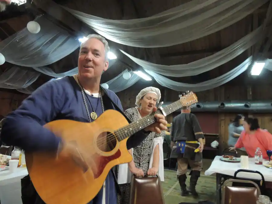 Ken Theriot smiles while playing an acoustic guitar at a festive indoor gathering with draped ceilings, joined by Lisa Theriot in period attire.
