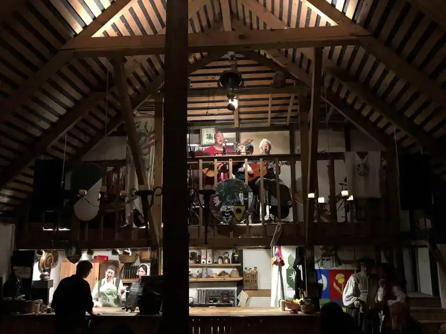 Ken Theriot performs with a band on the upper level of a rustic wooden hall during a medieval-themed gathering, while guests below socialize among banners and period decor.