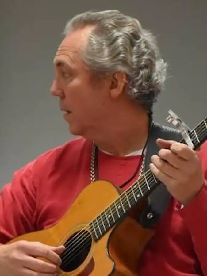 Ken Theriot wearing a red shirt and beaded necklace plays an acoustic guitar on stage, glancing to the side during his performance.