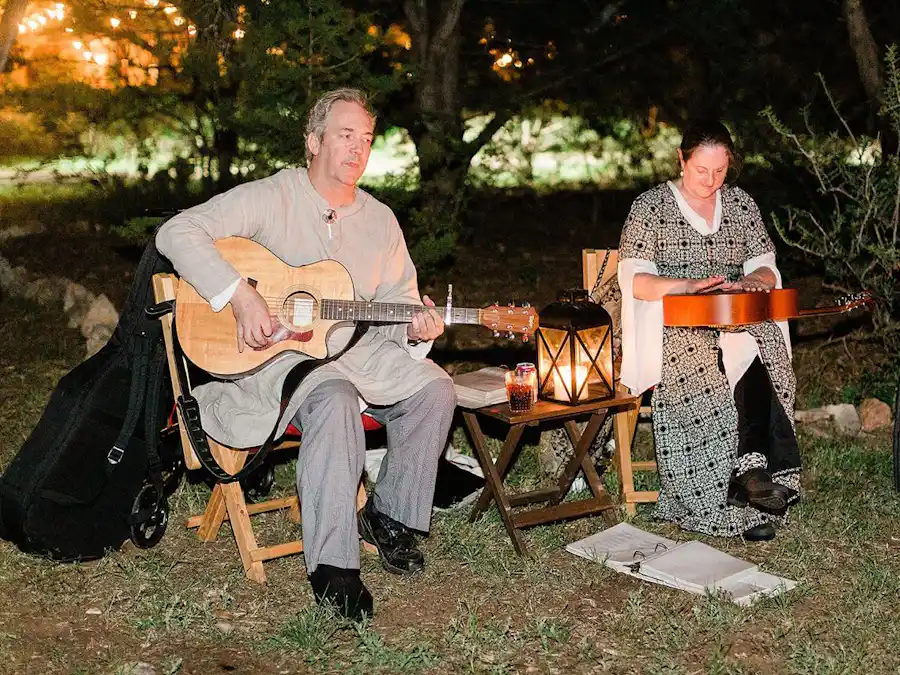 Ken Theriot sits outdoors at night playing an acoustic guitar beside Lisa Theriot, illuminated by a lantern during an intimate performance.
