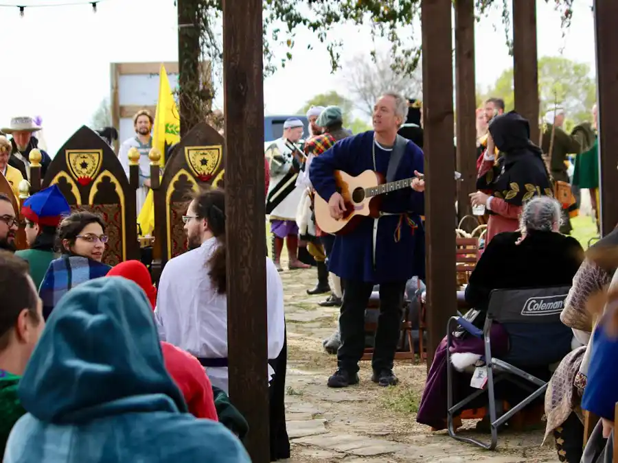 Ken Theriot plays guitar outdoors at a medieval-themed event, performing for a seated crowd amidst banners and costumed attendees.