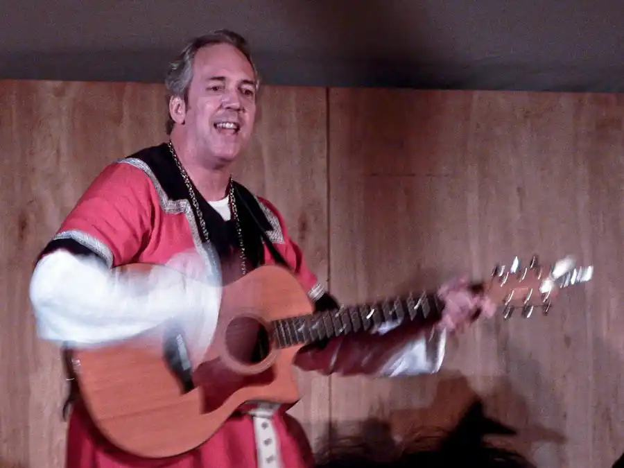 Ken Theriot smiles while performing songs in a red medieval tunic, strumming an acoustic guitar against a wooden backdrop.