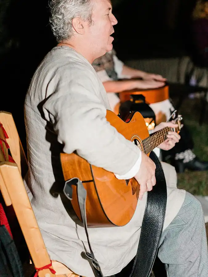 Ken Theriot in a beige tunic plays an acoustic guitar and sings during a nighttime performance, seated alongside another guitarist.