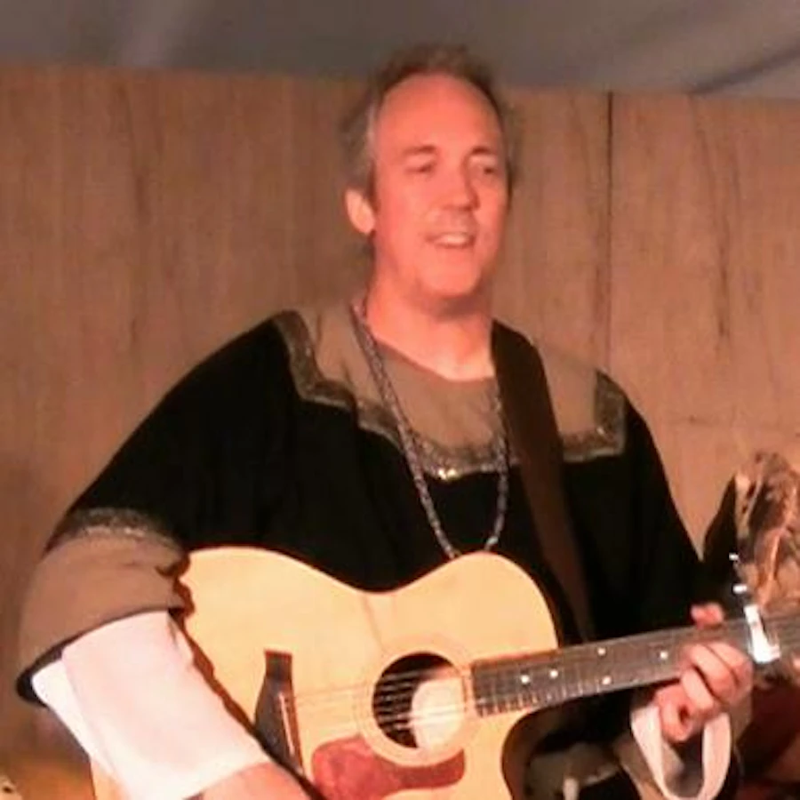 Ken Theriot dressed in medieval‑style attire plays an acoustic guitar on a wooden stage, smiling as he performs.