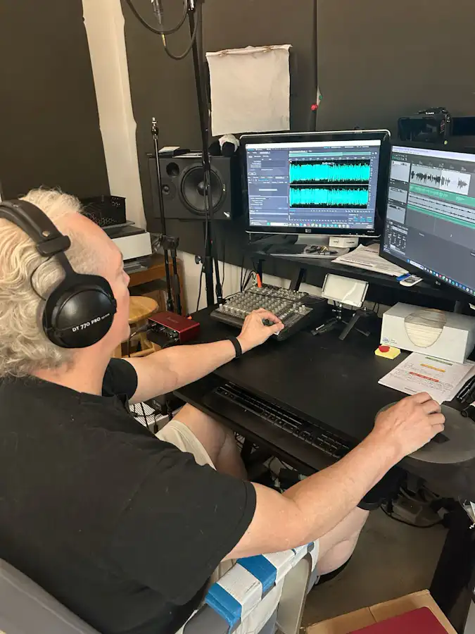 Ken Theriot wearing headphones sits at a digital audio workstation editing waveforms on dual computer screens with mixing equipment and speakers.