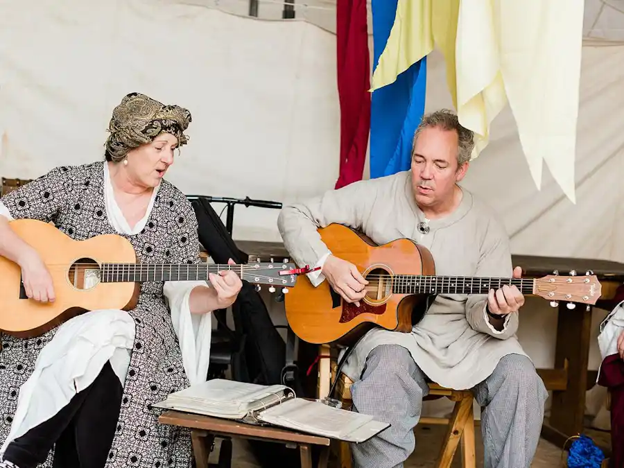 Ken Theriot sits in medieval attire playing an acoustic guitar beside Lisa Theriot, performing together with sheet music open on a stand in a costumed setting.
