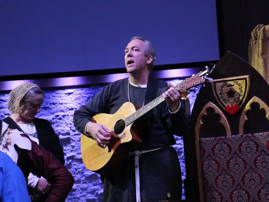 Ken Theriot performs on stage, singing and playing an acoustic guitar in medieval-style clothing with a fellow costumed performer beside him.
