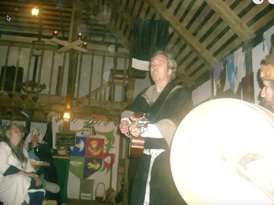 Ken Theriot wears a medieval costume and crown while playing guitar and singing in a rustic hall decorated with banners and onlookers.