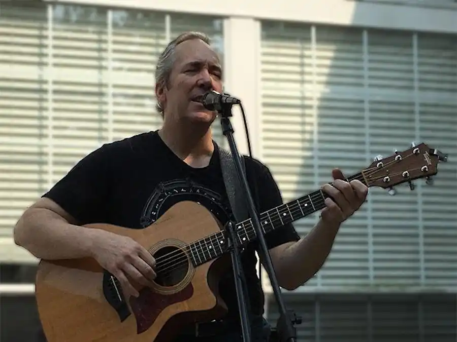 Ken Theriot sings into a microphone while playing an acoustic guitar during an outdoor performance.