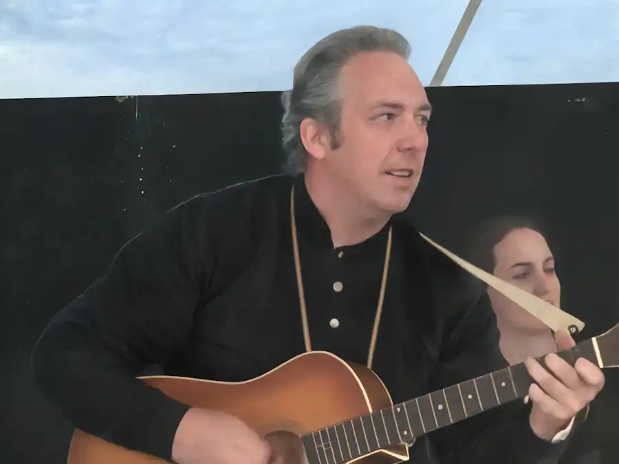 Ken Theriot strums an acoustic guitar with a fellow musician in the background during a live performance.